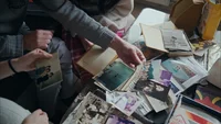 Mehrere Personen sortieren eine verstreute Sammlung alter Fotos, Briefe und Alben auf einem Tisch, wobei sie verschiedene Bilder in den Händen halten und untersuchen.