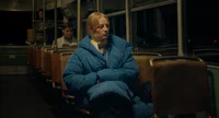 Eine Frau in einem blauen Mantel sitzt nachts allein in einem fast leeren Bus und blickt mit nachdenklicher Miene hinaus. Eine andere Person sitzt weiter hinter ihr und liest ein Buch. Das Innere des Busses ist nur schwach beleuchtet.