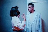 Eine Frau mit Brille und Maske gestikuliert im Gespräch mit einem ernst dreinblickenden Mann in einem hellen Blazer, der in einem schwach beleuchteten Raum vor einer weißen Tür steht.